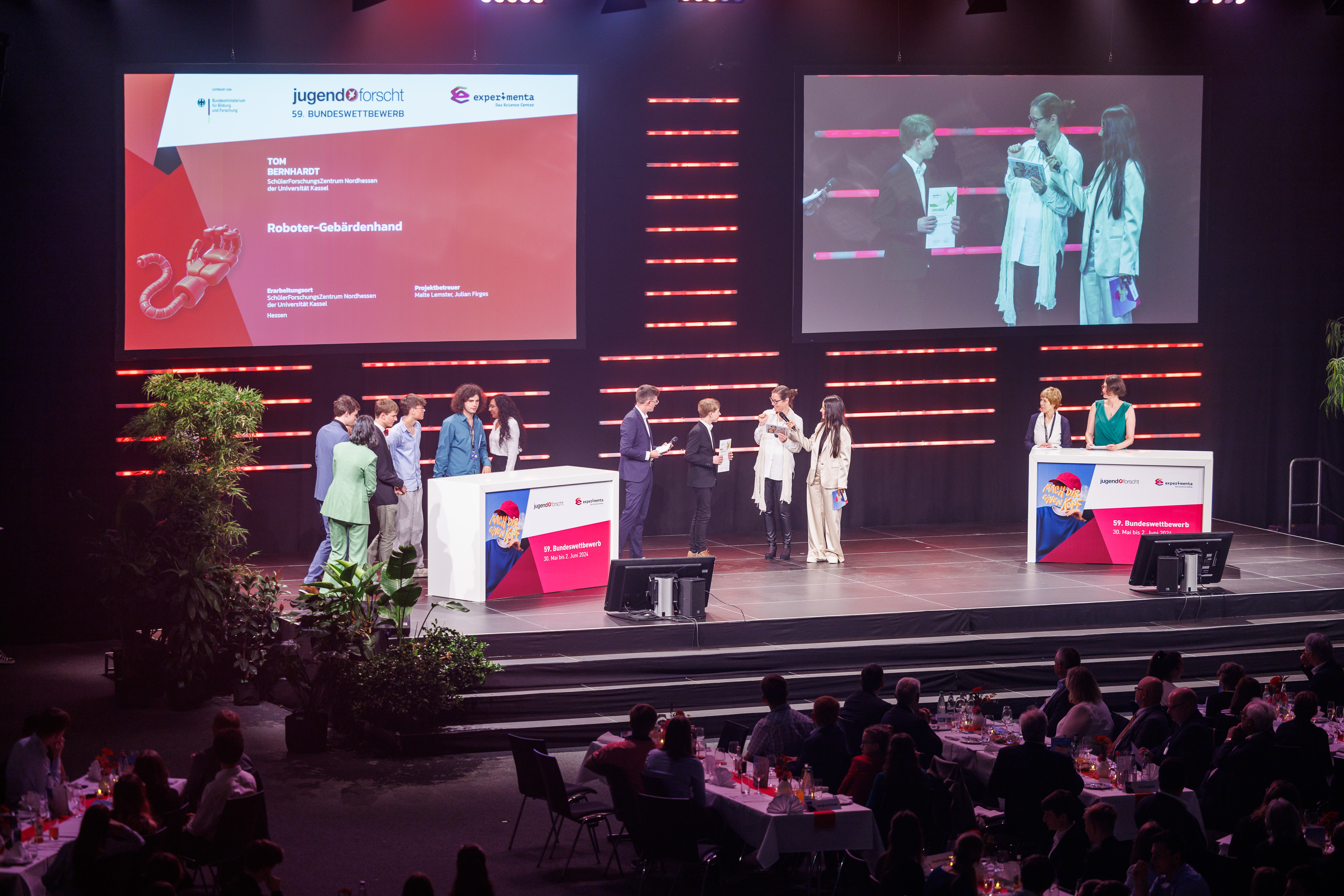 Preisverleihung: Preisträger stehen auf der Bühne mit ihren Urkunden. Im Vordergrund sitzt das Publikum, im Hintergrund ist großer Screen mit den Namen und ein Screen mit einer Livekamera zu sehen.